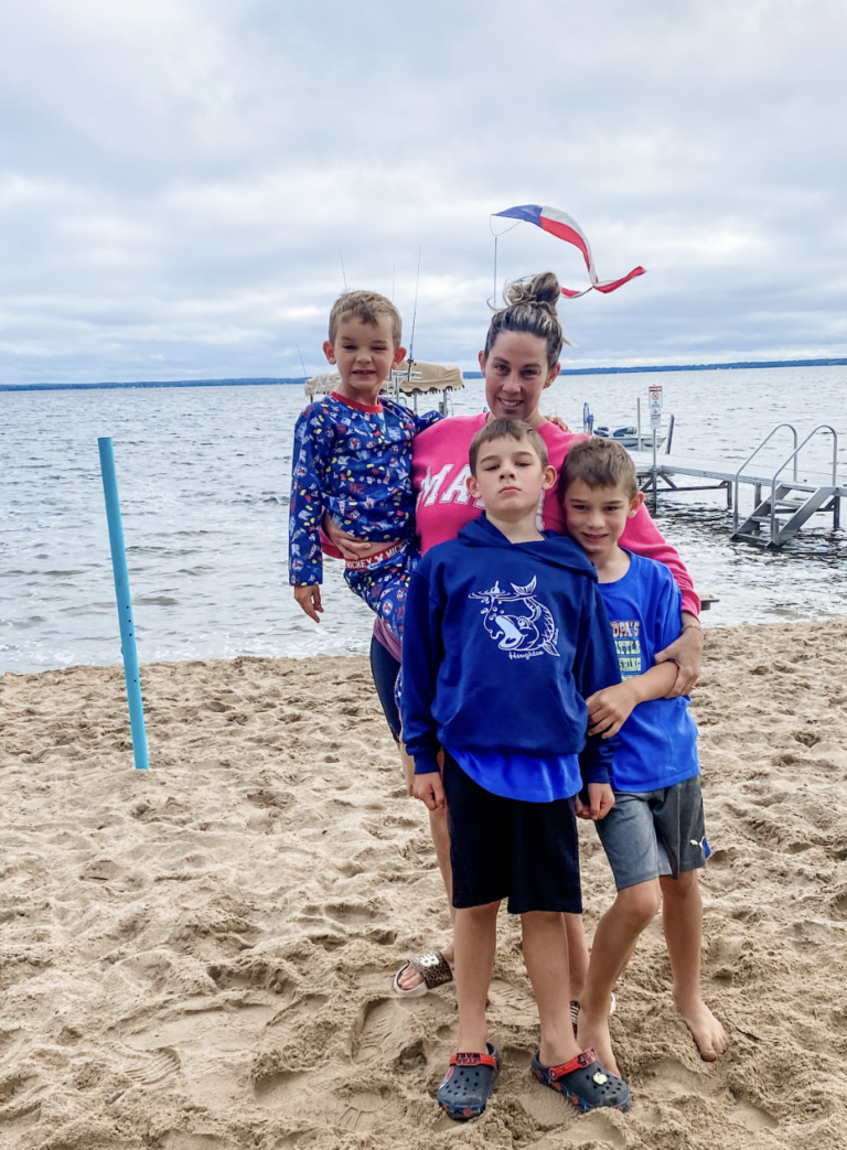 A mom and her three sons embrace on the sand in front of a dock.