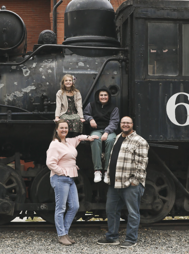 A man and woman pose in front of a steam engine train with their son and daughter.
