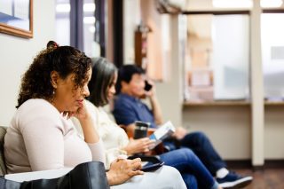 People sit in doctor's waiting room. One woman looks a medical brochure and a man uses his smart phone to make a call.